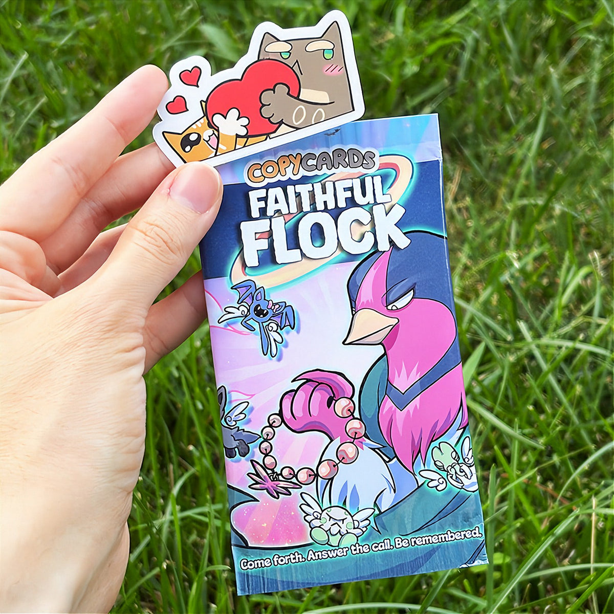 A hand holds a cartoon sticker next to a printed card pack titled "CopyCards Faithful Flock" on grass outdoors.