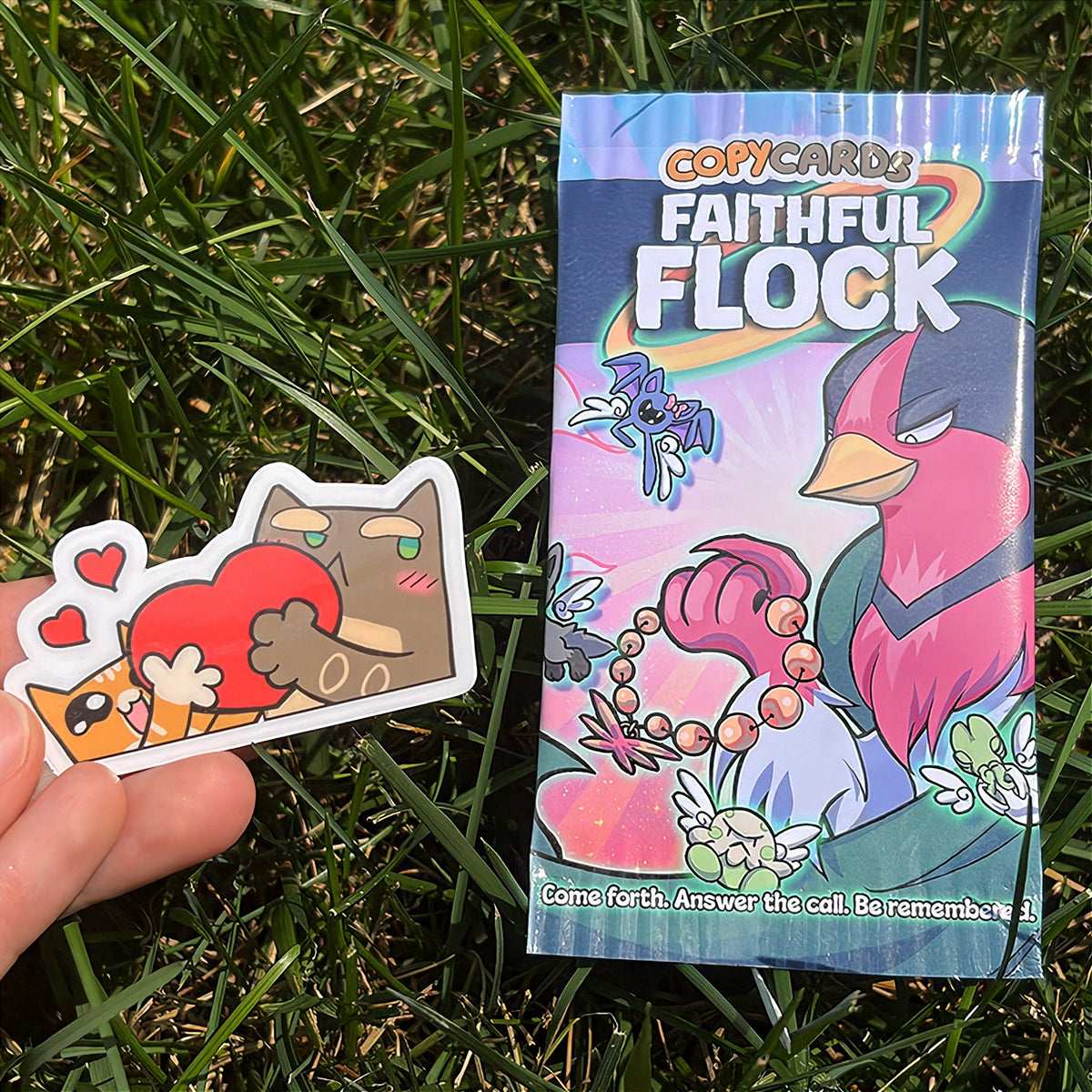 A hand holds a cartoon sticker next to a printed card pack titled "CopyCards Faithful Flock" on grass outdoors.