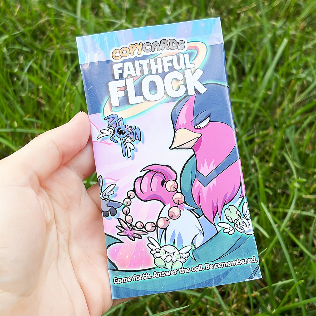 A hand holds a cartoon sticker next to a printed card pack titled "CopyCards Faithful Flock" on grass outdoors.